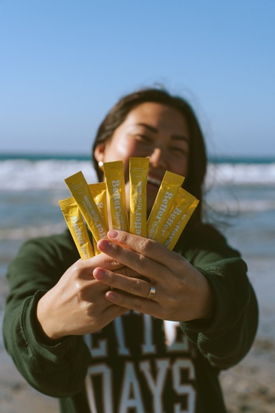A high-energy lifestyle image showing a person holding multiple hydration supplement sachets against a bright ocean background.
